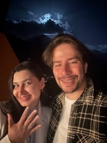 Bruno e Luciana sorrindo à noite; Luciana mostra a mão com um anel