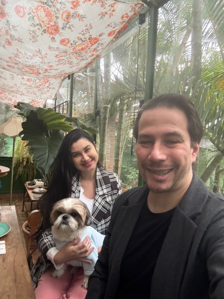 Bruno e Luciana sorrindo à mesa com o cachorro no colo, entre plantas